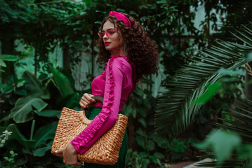 Fashionable curly woman wearing pink ruffled blouse, headband, sunglasses, holding straw wicker top handle bag, posing in tropical garden. Summer fashion conception. Copy, empty space for text  - obrazy, fototapety, plakaty