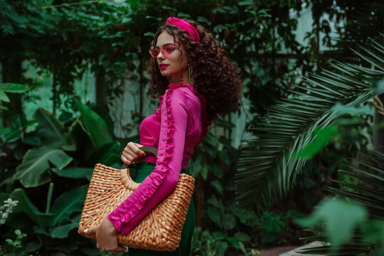 Fashionable curly woman wearing pink ruffled blouse, headband, sunglasses, holding straw wicker top handle bag, posing in tropical garden. Summer fashion conception. Copy, empty space for text 