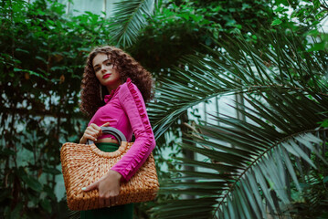 Fashionable curly woman wearing pink asymmetric ruffled top,  holding straw wicker top handle bag,...