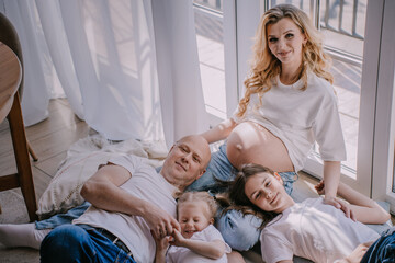 Happy swedish family in white t-shirts and blue jeans laying on the floor, smiling. Caucasian family at home on weekend. Pregnant woman with family at home on sunny day.