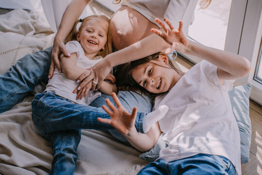 Cropped shoot of two cheerfull sisters in blue jeans and white t-shirts laying on the floor close to pregnant moms belly looking at camera. Happy family at home playing together. Pregnancy concept.