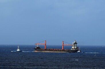 Large cargo ship with cranes being pulled by two blue and white tugboat, blue ocean with blue sky in the background