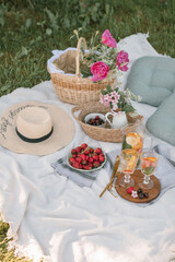 Strawberries in bowl, lemonade, straw hat, peony flowers, pillows. on white blanket. Outdoor gathering concept. Beautiful plastic free picnic outdoors.