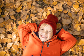 Child in an orange jacket lies on the fallen leaves in the autumn forest. Rest in the fall in...