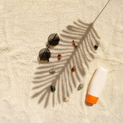 Fototapeta na wymiar palm leaf shadow of natural sunlight on sandy color towel with sea rocks and seashell, sun cream and sunglasses. Summer vacation concept, square composition with copy space.