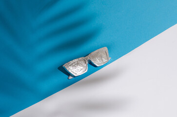 Sunglasses wrapped in foil on a pastel blue and beige background. Tropical summer concept. Minimal composition.