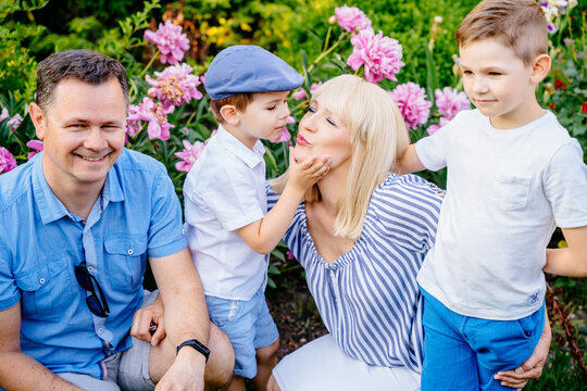 Portrait of family, caucasian husband his blond charming wife hugging with their cute two little sons in garden or park with peonies on background. Little boy kiss mom.