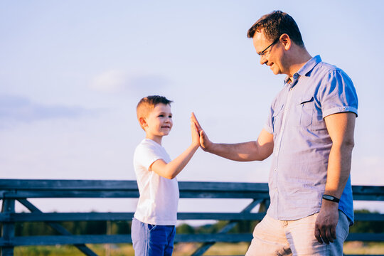 School Age Boy And His Dad Doing High Five And Feeling Wonderful Outdoor At Summer Vacation.