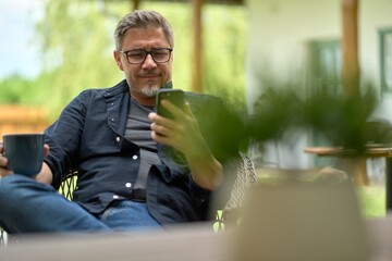 Portrait of middle aged man at home outdoor in garden, sitting on terrace, relaxing, drinking coffee, reading news on phone.