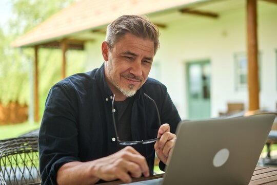 Middle Aged Man Working With Laptop Computer At Home Outdoor In Garden. Businessman In Home Office, Online, Thinking.