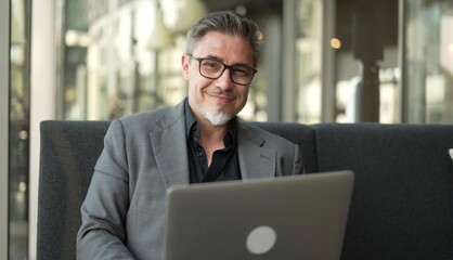 Businessman sitting on couch working with laptop computer in office lobby. Portrait of happy middle aged man in business casual, smiling.