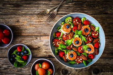 Fresh salad - goat cheese, raspberries, blueberries, pecan nuts, cherry tomatoes, pickled radishes and leafy greens on wooden table
