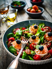 Fresh salad - goat cheese, raspberries, blueberries, pecan nuts, cherry tomatoes, pickled radishes and leafy greens on wooden table
