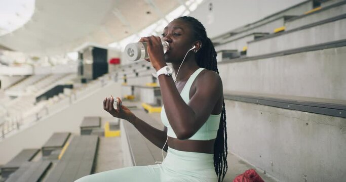 Young athlete listening to music, hydrating and getting ready to train at a stadium. Sporty black woman enjoying her favourite songs before a workout. Fit girl preparing to exercise at a sports club