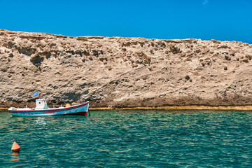 Fototapeta premium Anchored small fishing boat under Greek flag, azure sea waters, clear blue sky, rocky hill, red fishing buoy floating. Frontal shot. Milos, Greece.