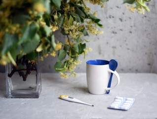 On the table next to a bouquet of linden blossom branches, a glass of linden tea, a thermometer and pills on a gray background.