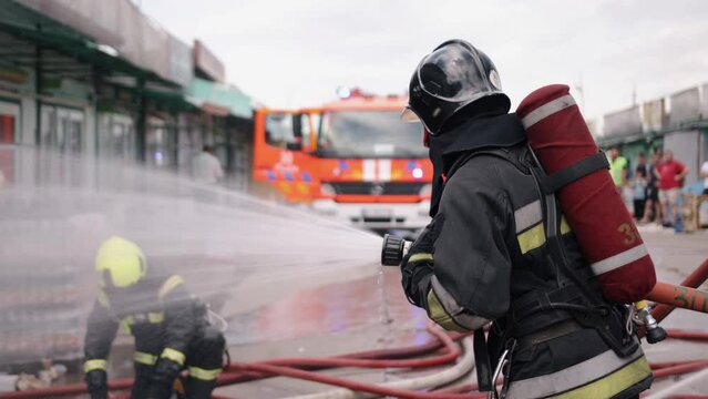 A firefighter is fighting a fire, a fire truck is in the background, and water is coming out of the hose