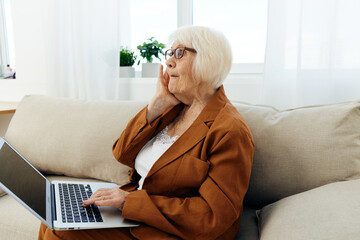 a sad elderly woman with problems at work is sitting at home on the sofa opposite the window and holding her hand near her face in a frustrated gesture looks at the laptop monitor