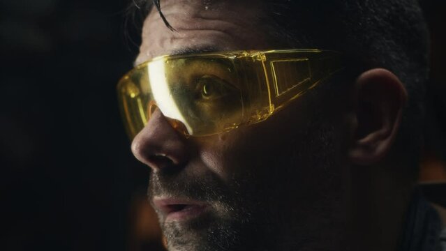 Exhausted worker in protective goggles and gloves wiping sweat from forehead after processing metal products for his small business in a dim garage