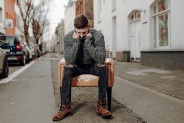 ashamed man in thoughts sit on old grungy chair in the middle of a street of a winter cold city and...