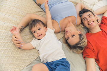 Cute family of three - mother, father, and their son - laying down together on a bed on their backs, laughing, enjoying their free time. High quality photo