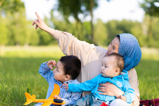 asian family resting in park, looking at sky. boy playing with toy plane. Muslim mother in hijab show to Kazakh child pilot flying airplane. islamic kid aviator dreaming, traveling. Startup, freedom