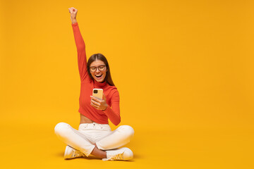 Excited happy woman sitting on floor with phone, shouting yes as winner, isolated on yellow