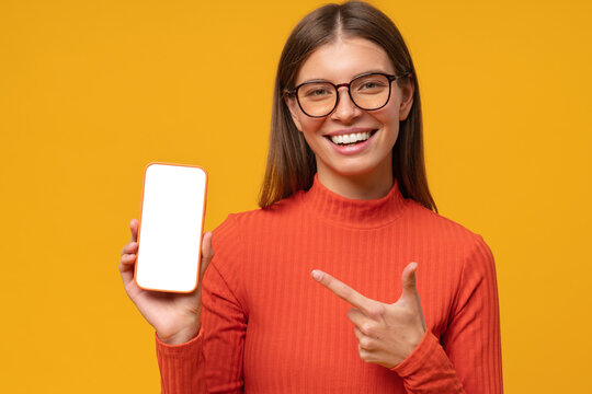 Portrait of happy young woman pointing to blank phone screen, isolated on yellow background