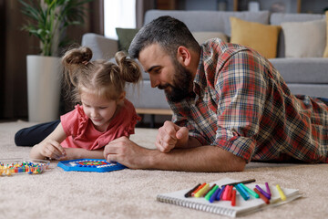 Fototapeta premium Young handsome father and cute daughter playing together. Dad and child making a puzzle with mosaic pegboard lying on the carpet at home. Educational toys for children. Home leisure. Father's day
