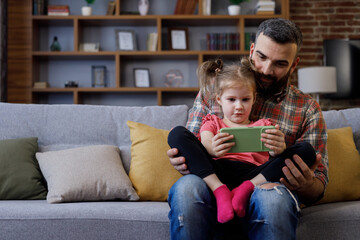 Happy cheerfull family. Young father and cute daughter watching funny videos or playing games on smartphone. Dad with little child girl enjoy spending time together, home leisure. Father's day