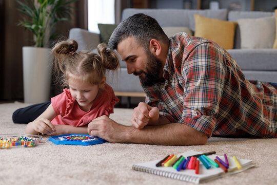 Young handsome father and cute daughter playing together. Dad and child making a puzzle with mosaic pegboard lying on the carpet at home. Educational toys for children. Home leisure. Father's day