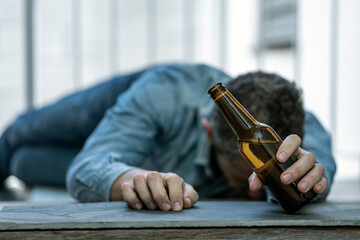 DRUNK MAN LYING ON THE FLOOR IN THE STREET ASLEEP WITH A BOTTLE OF BEER IN HIS HAND. ALCOHOL CONSUMPTION ADDICTION. ALCOHOLISM CONCEPT. FOCUS SELECTED.