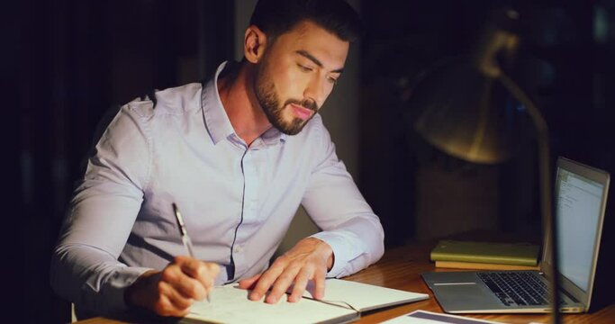 Young business man working overtime and taking notes writing in a diary while working on a laptop in an office late at night. Male professional reading an online search result and planning in a book