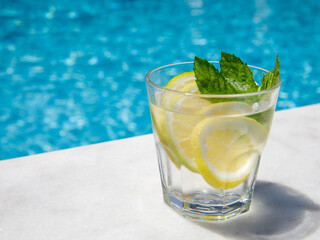 Citrus and mint leaves infused water, cocktail, lemonade in glass near pool. Summer iced cold drink with lemon, lime, ice and mint on blue water and white marble background, copy space, side view