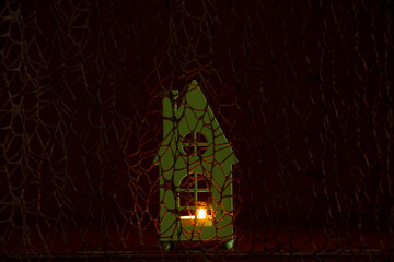 A small children's toy house with a candle in the window against the background of a grid of fabric in the dark, light in the window of the house