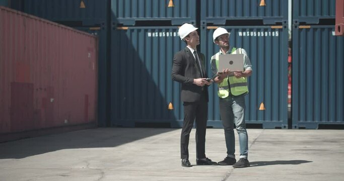 Foreman in hardhat, safety vest and glasses using laptop to explain about containers data to Asian businessman. They talking and prepare loading container box at warehouse logistic in cargo freight.