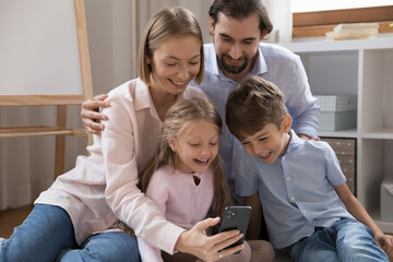 Happy joyful couple of parents and two little kids making video call on mobile phone, chatting together, taking family selfie, resting on warm floor at home, looking at screen, webcam, laughing