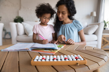 Caring African mother helps with maths task to little cute daughter, close up view abacus on table. Children learning, preparation for school, mathematics class with tutor at home, education concept