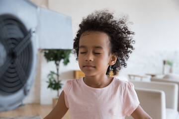 Close up tranquil little African girl stand in front of blowing fan closes eyes enjoy fresh air in...