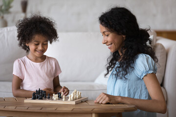 In living room African mother engaged in board game with cute little daughter, family enjoy chess...