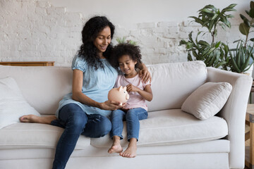 Young African mum and little daughter sit on sofa drop coins cash into piggybank, save money,...