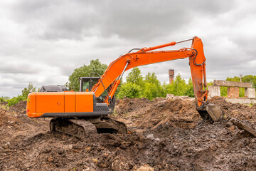 Orange excavator digs trench in ground against backdrop of green trees and blue summer sky.