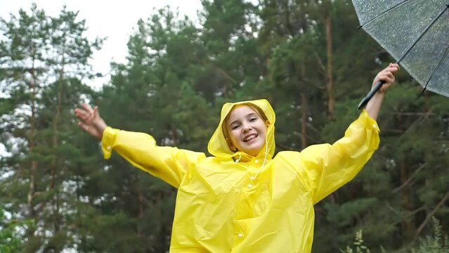 Cheerful girl in yellow raincoat jumps high spreading hands to sides. Cute teenager has fun in forest on rainy day holding umbrella slow motion