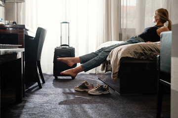 woman taking off footwear in sunny hotel room on the bed. Tourist relaxing on hotel room after travelling with suitcase. Happy female having rest after long trip with language Dark silhouette 