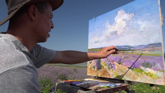 A male artist draws a picture on canvas outdoors using a palette knife. Plein air oil painting. Rural landscape, blooming lavender fields