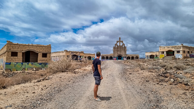Tourist man looking at abandoned leper village of Sanatorio de Abona near Abades beach, east coast Tenerife, Canary Islands, Spain. Dirt hiking trail leading to concrete church in center of ghost town