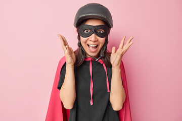 Emotional superhero wears helmet mask and cape keeps hands raised and exclaims loudly reacts to awesome news prepares for costume party isolated over pink background. Masquerade is about to start