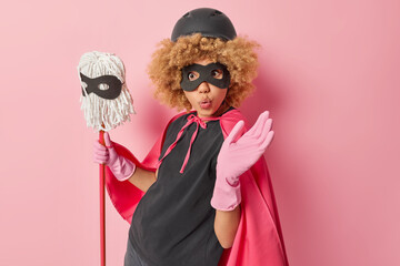 Surprised woman pretends being superhero does housework washes floor with mop supplies cleanliness has wondered expression isolated over pink background. Daywork routine and housekeeping concept