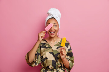Horizontal shot of cheerful woman poses with gourment tasty ice cream has playful mood wears domestic clothes towel after taking shower has fun looks away isolated over pink background. Summer time