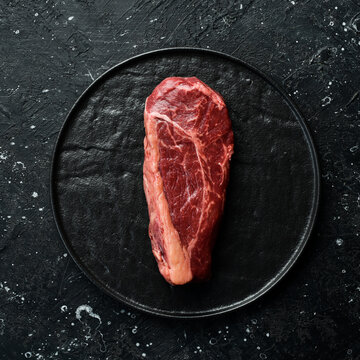 Classic raw New York beef steak. Fresh meat with seasonings. Aged steak. Top view. On a black stone background.